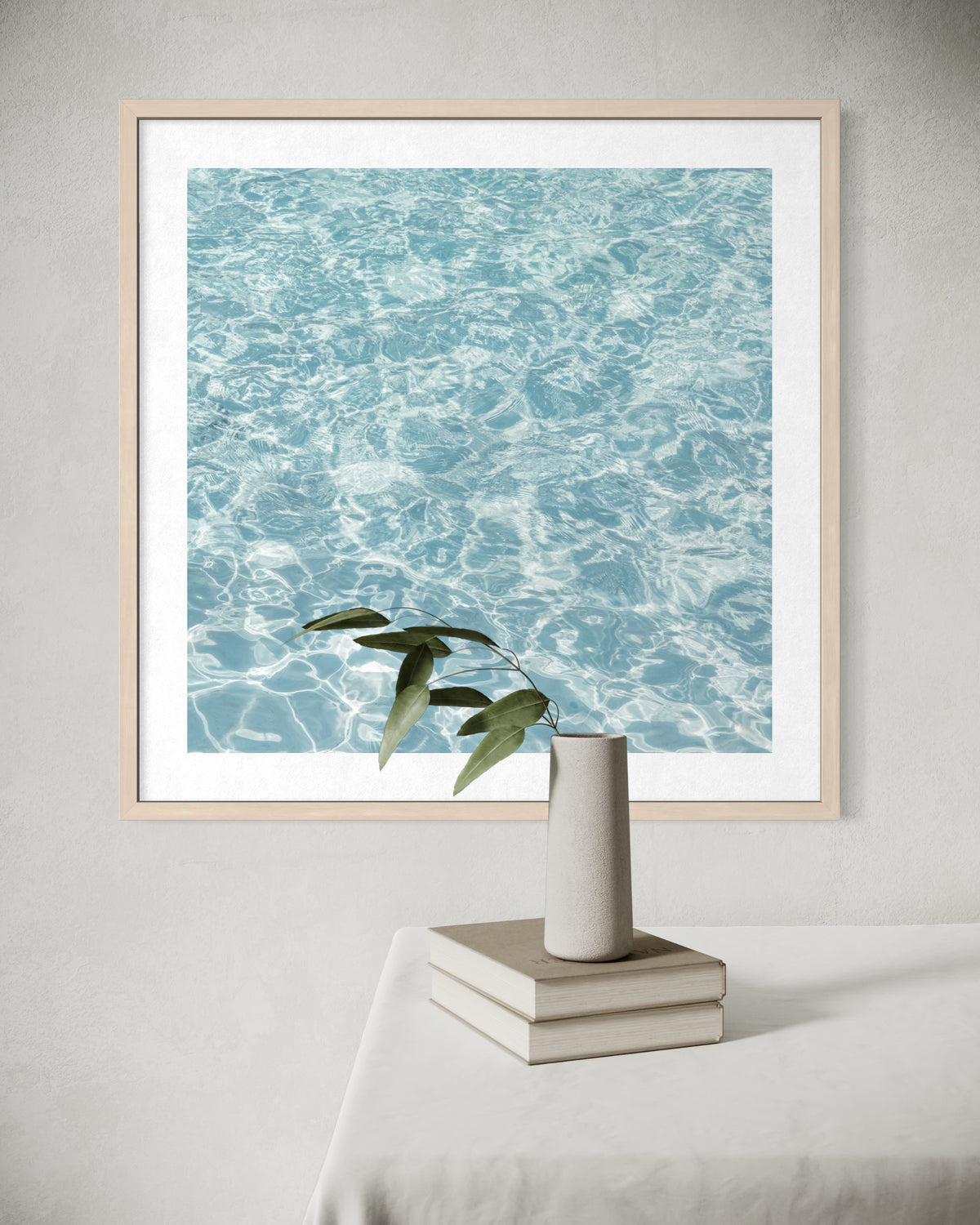 Framed print of calm water titled “Refresh” displayed above a table with stacked books and a glass vase.