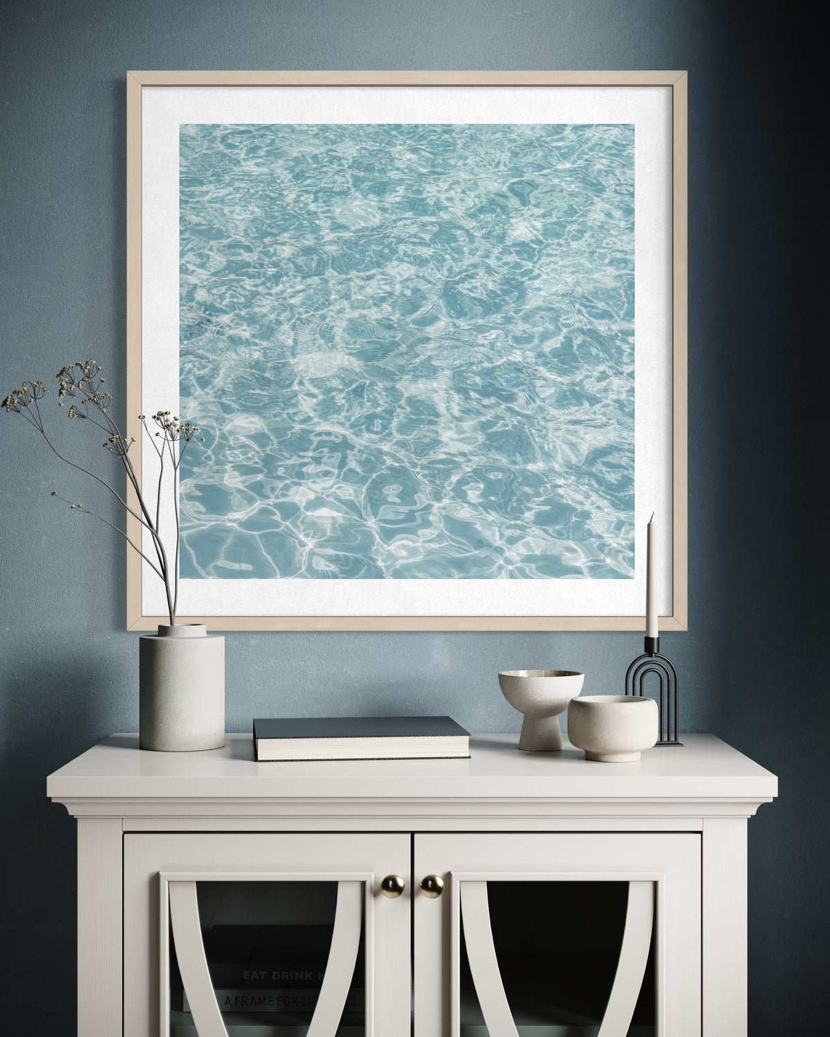 Framed print of calm water titled “Refresh” hangs above a white cabinet on a blue wall with minimal decor.