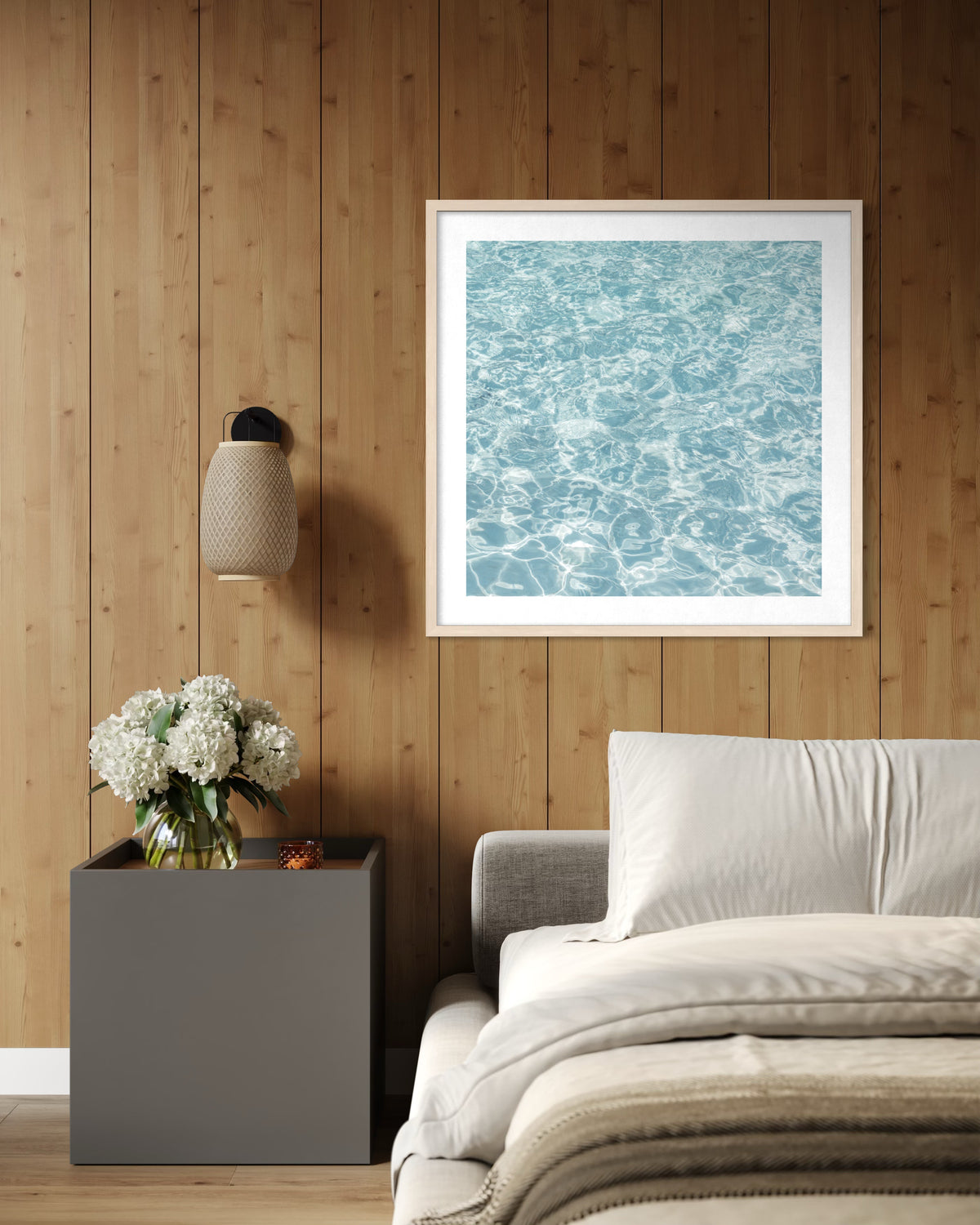 Bedroom with light panel walls, white bedding, water artwork, a hanging lamp, and a vase of white flowers.