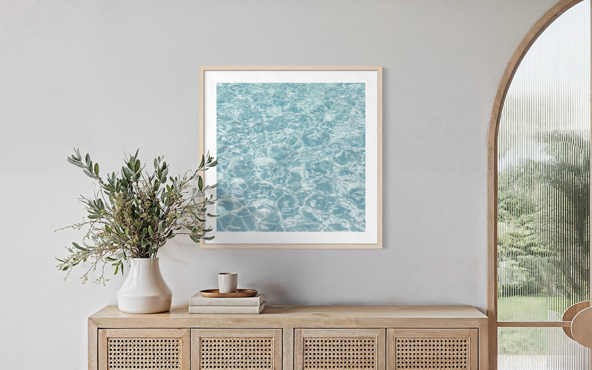 Framed print of tranquil water scene above a wood sideboard, vase with greenery, and stacked books.