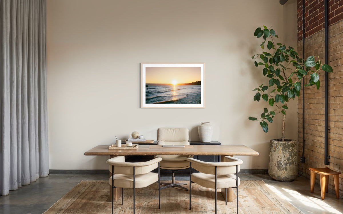 Framed “Surfers at Sunset” photo on wall above wooden desk with two chairs and a large indoor plant.