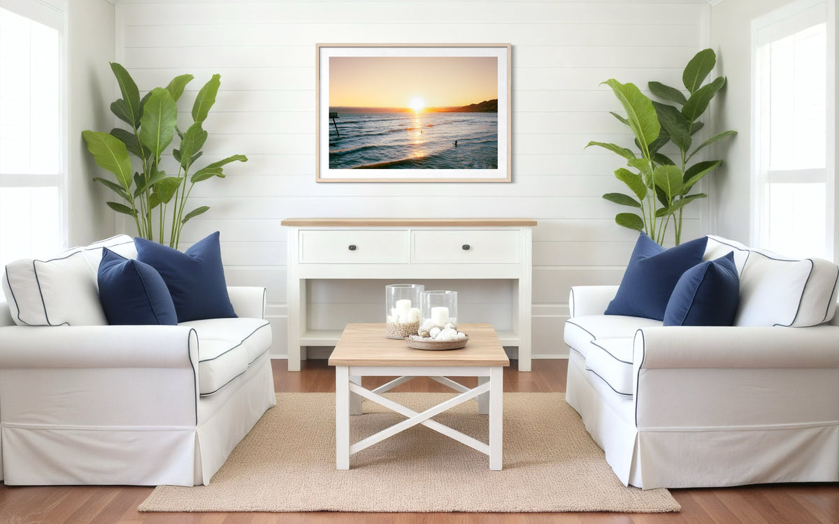 White sofa with blue pillows, plants, and a framed Surfers at Sunset print on the wall in a bright living room.