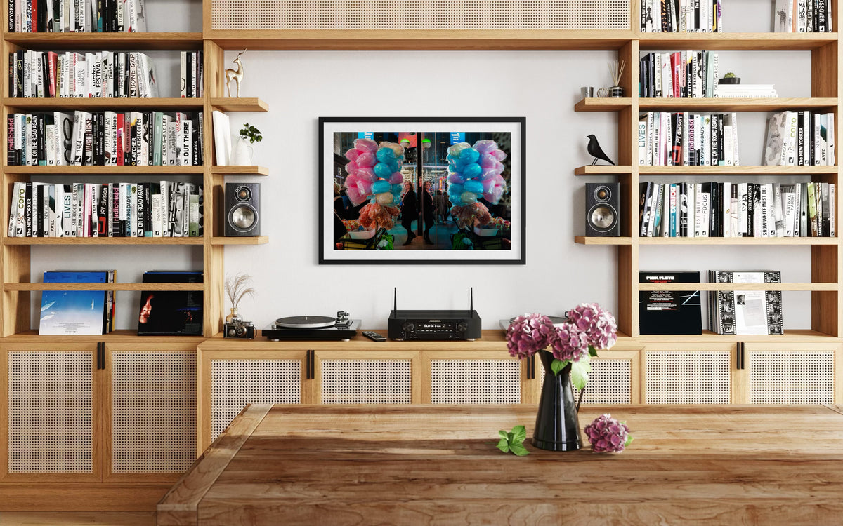 Framed “Sweet Reflections” photograph above shelves with books and a vase in a bright living room.