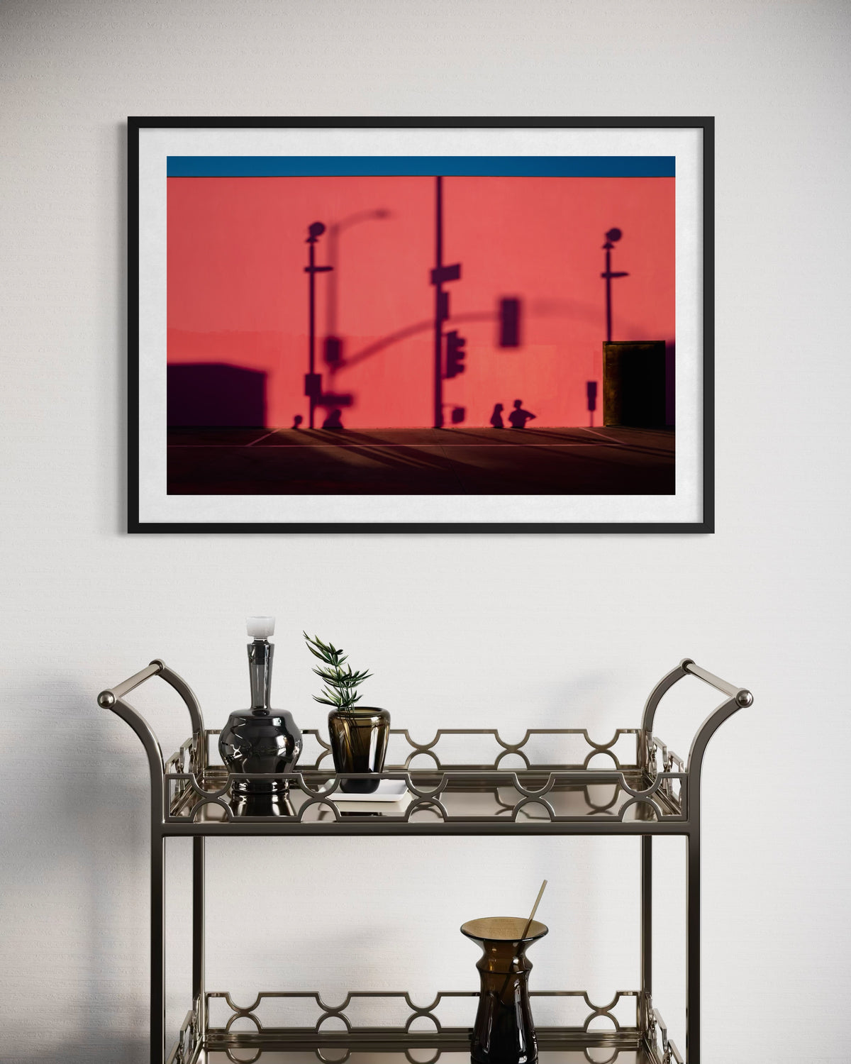 Framed print of city shadow patterns on a pink facade, captured above a bar cart with bottles, glasses, and a plant.