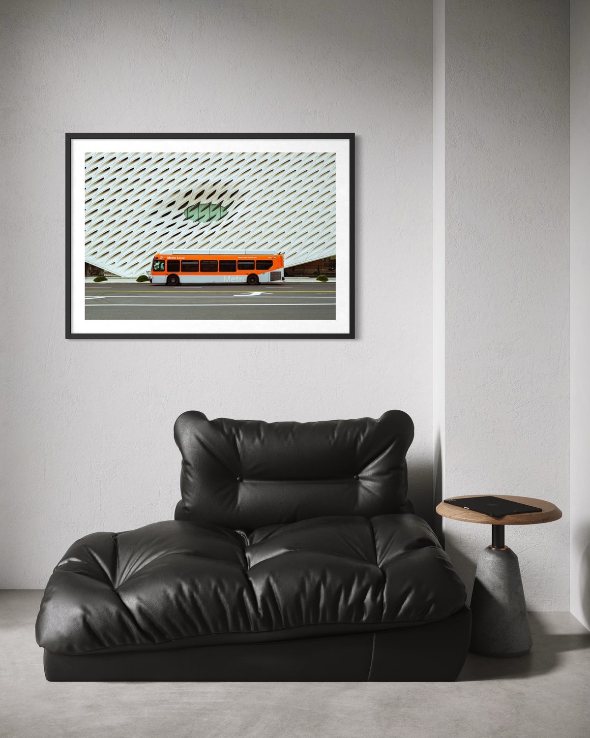 Metro bus drives through downtown Los Angeles, seen in a framed photo above a chair and side table against a white wall.