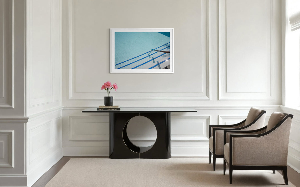 Framed “The Hotel Pool” photo hangs above a black console, beige chairs, and pink flowers in a minimalist room.
