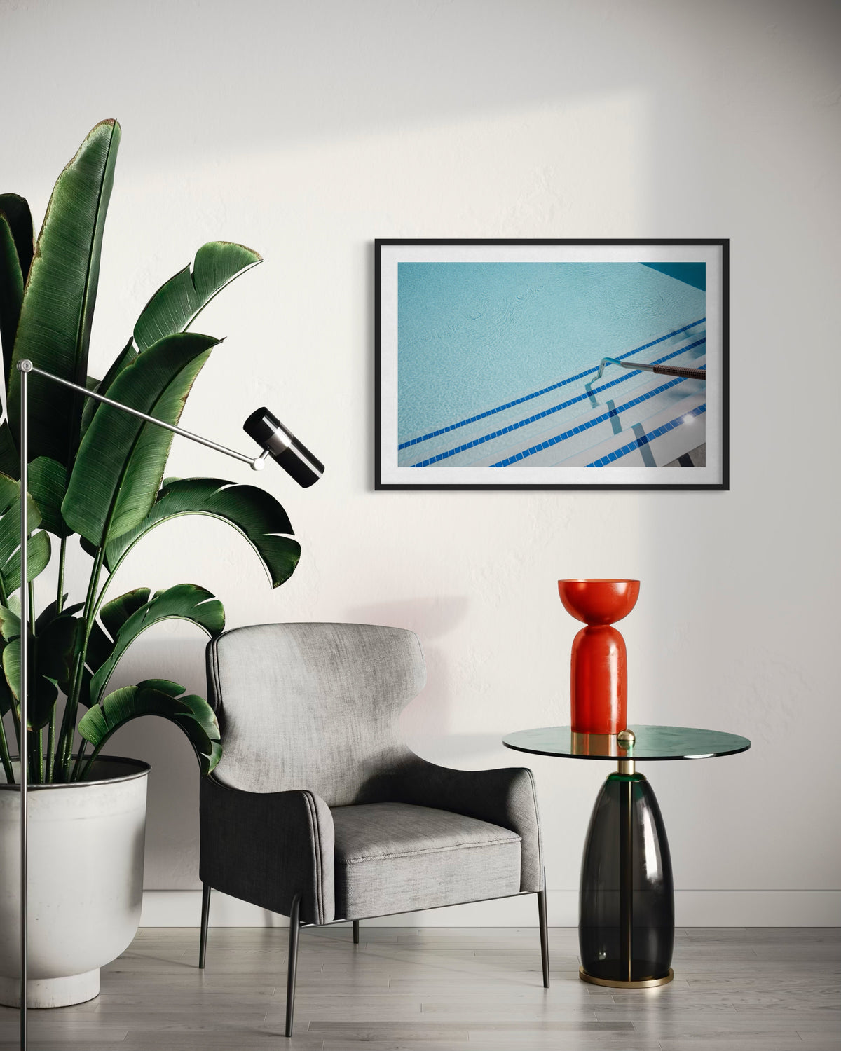 Framed “The Hotel Pool” photo on white wall above a gray armchair, side table with red vase, and a large plant.
