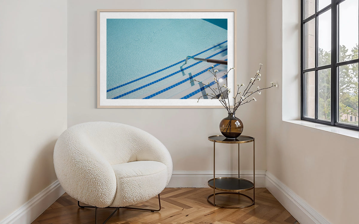 Framed “The Hotel Pool” photograph of a pool scene on wall above light chair and table in modern room.