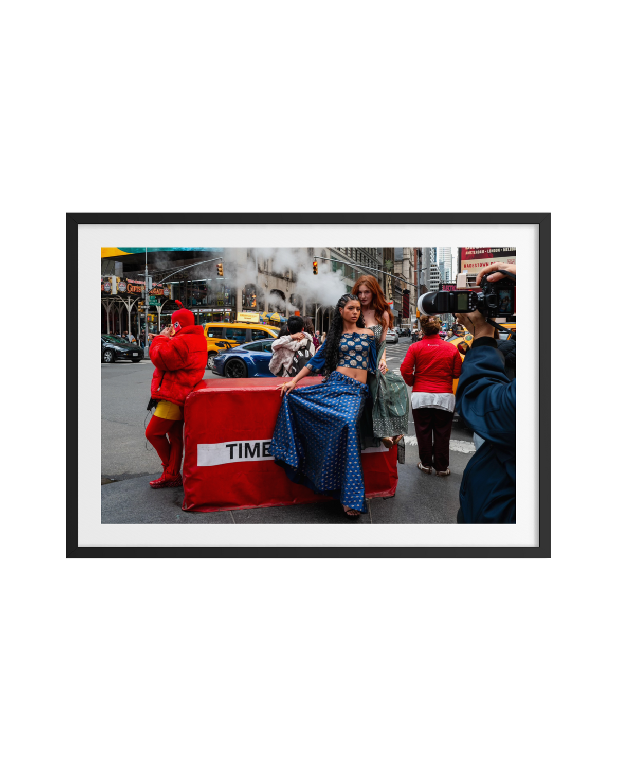 Two women sit on a red barrier in Times Square, New York, with a yellow taxi and pedestrians nearby.