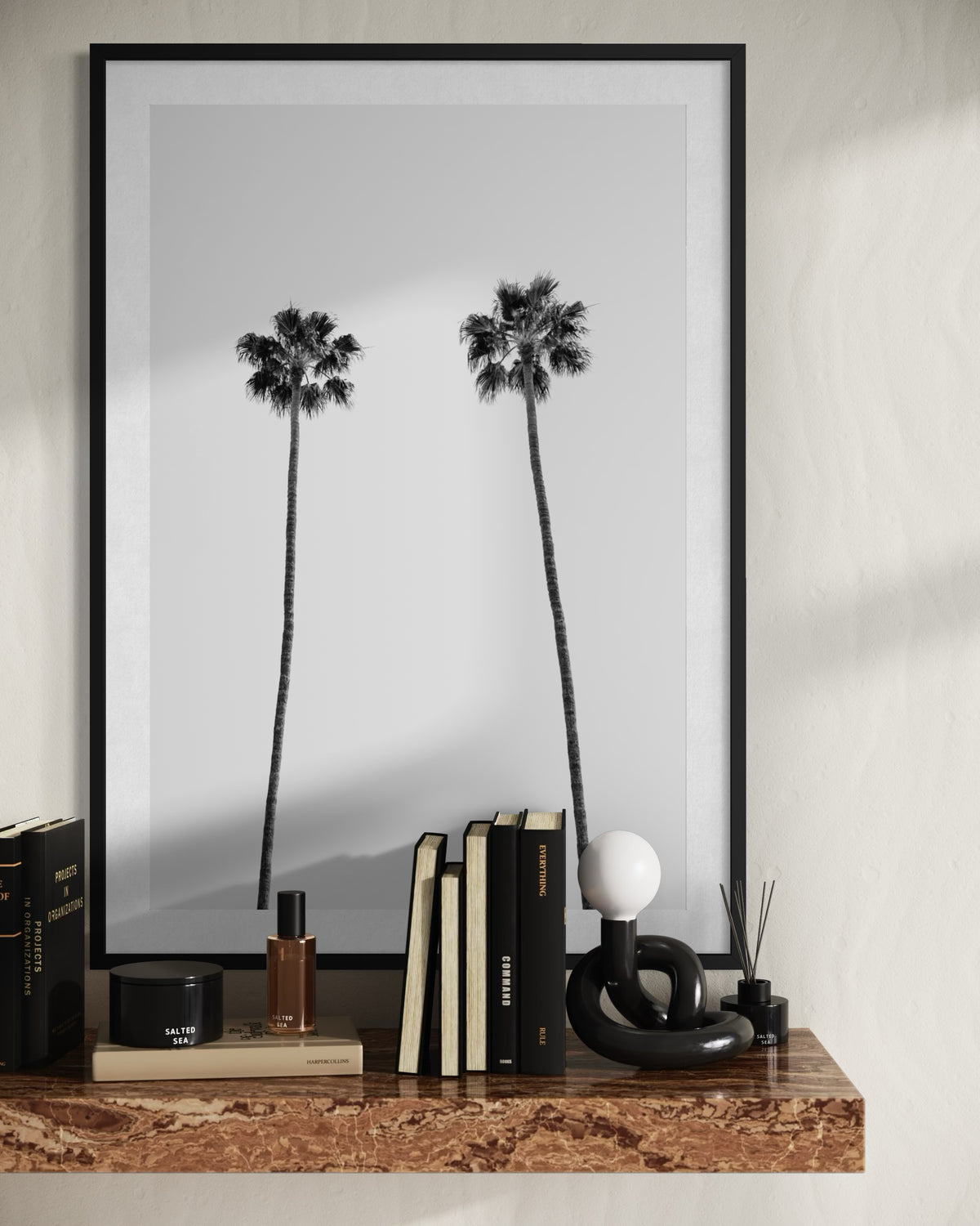 Black and white photo of two tall palm trees in Los Angeles, shown above a shelf with books, perfume, and a lamp.
