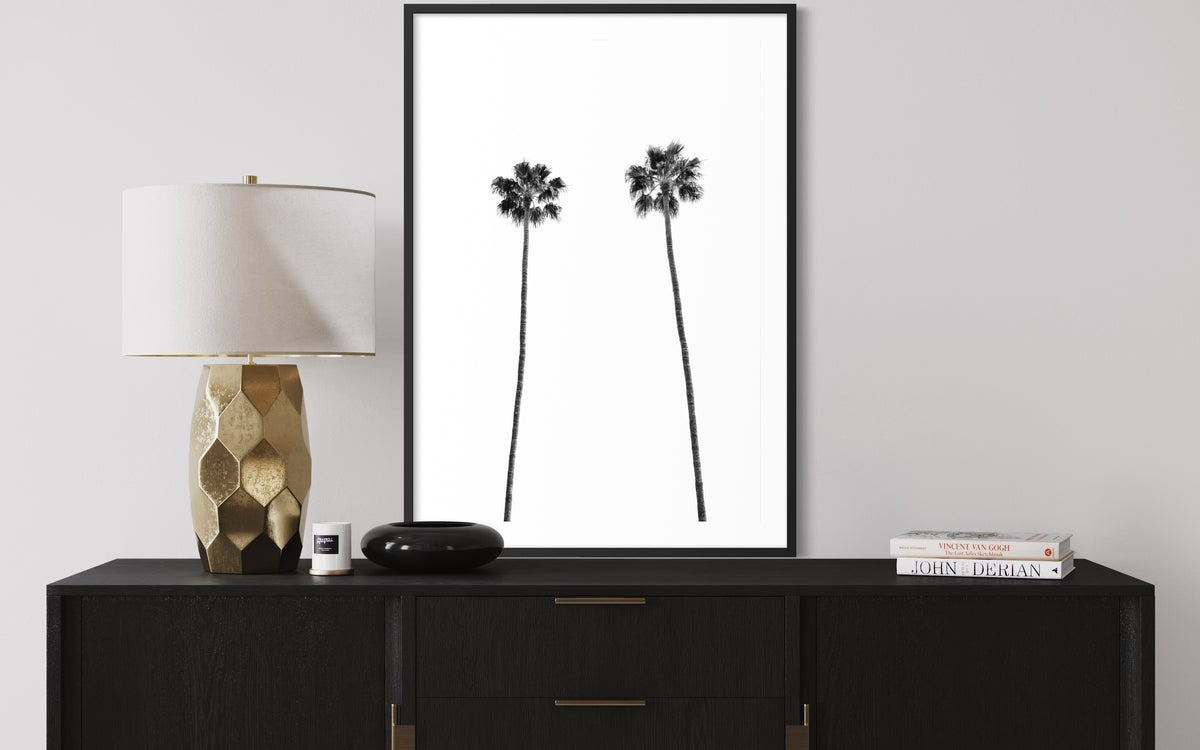Framed Twin Palms photograph above a black dresser with gold lamp, bowl, and books in a minimal room setting.