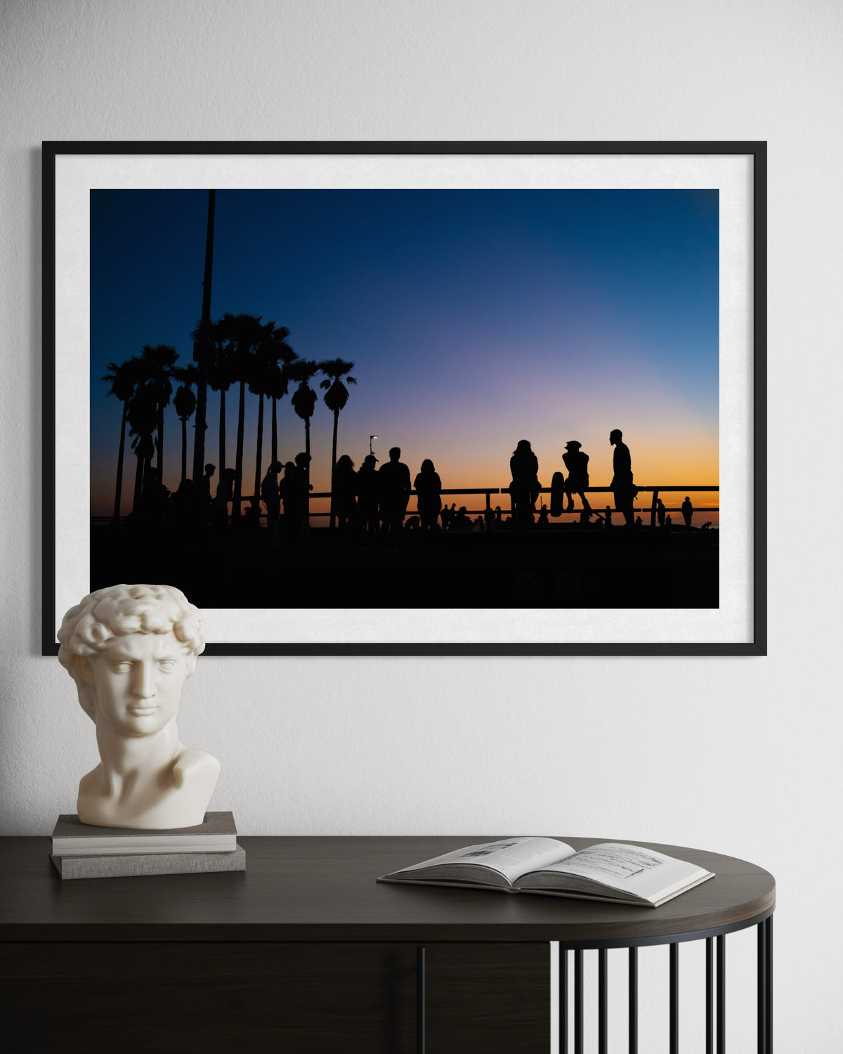 Framed print of silhouetted palm trees against a Venice Beach sunset, displayed above a table with decor.