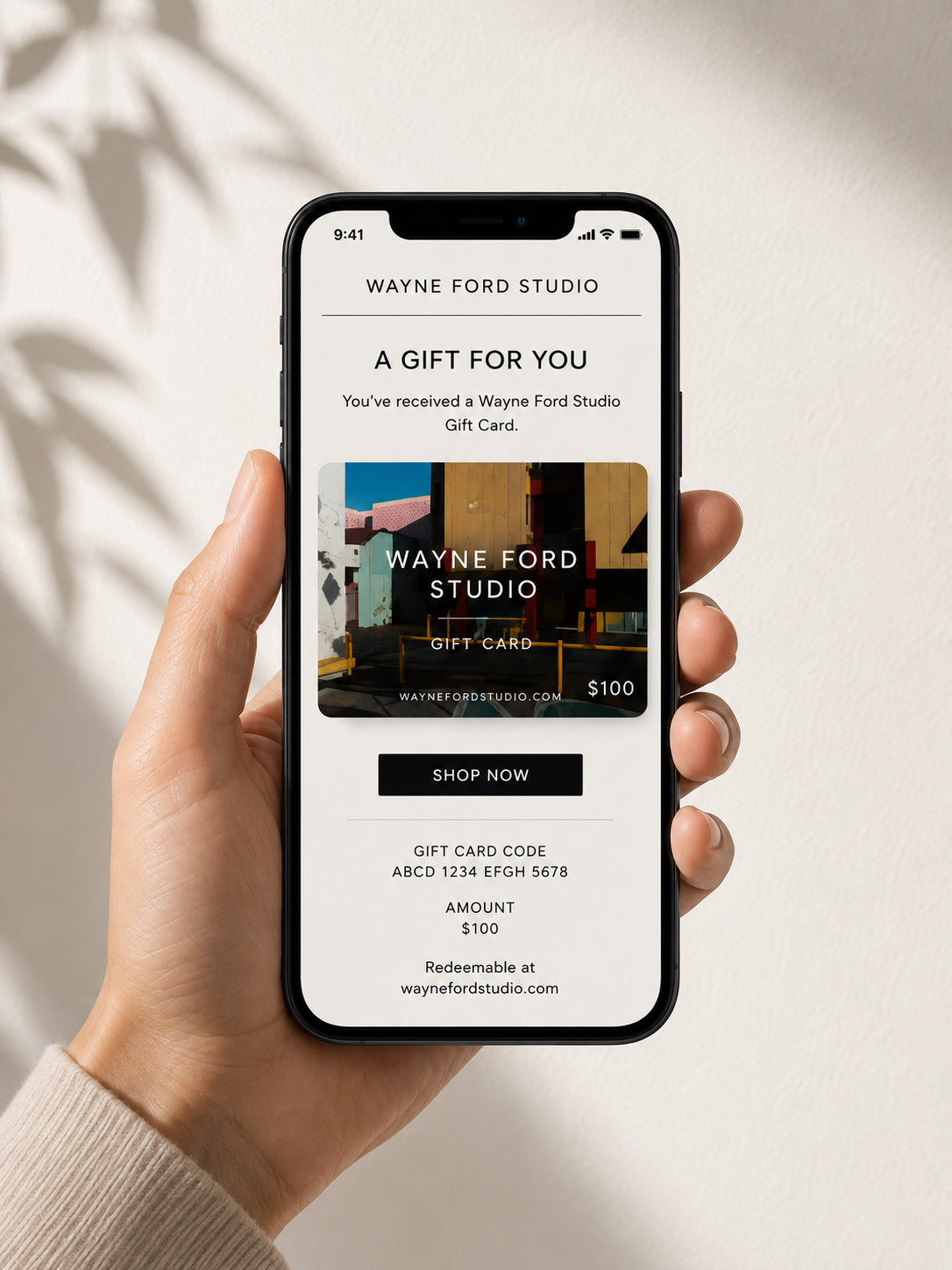 Hand holding a phone displaying a $100 Wayne Ford Studio Gift Card, vibrant abstract background behind.