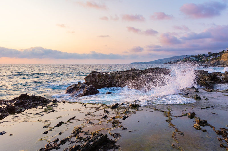 Waves crash against rocks at sunset on Laguna Beach, California under a partly cloudy summer sky.