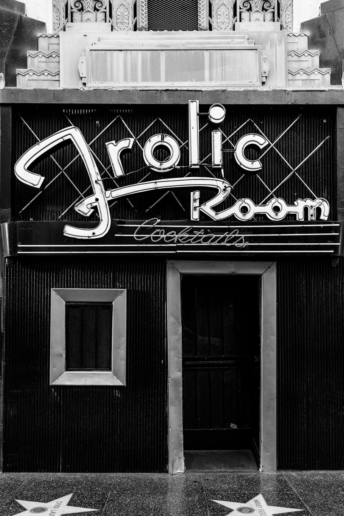 Black-and-white view of the Frolic Room entrance and neon signs along Hollywood Boulevard’s Walk of Fame in Los Angeles.
