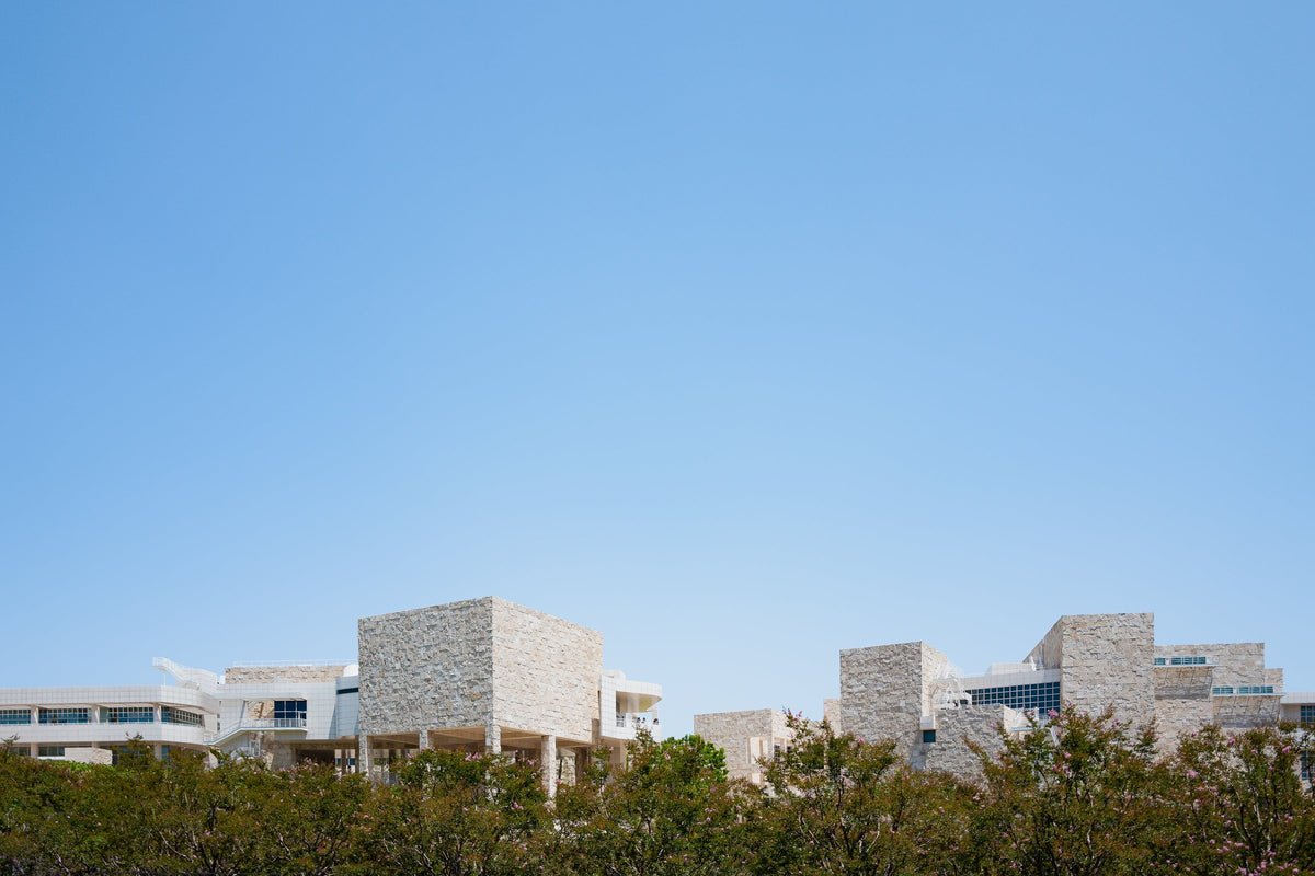 Travertine buildings of Getty Center III in Los Angeles rise above green trees under a clear blue sky.