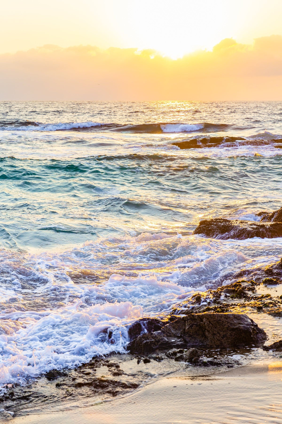 Golden sunlight reflects on ocean waves hitting a rocky shore at sunrise.
